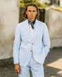 Man wearing a light blue striped suit with a patterned scarf outdoors.