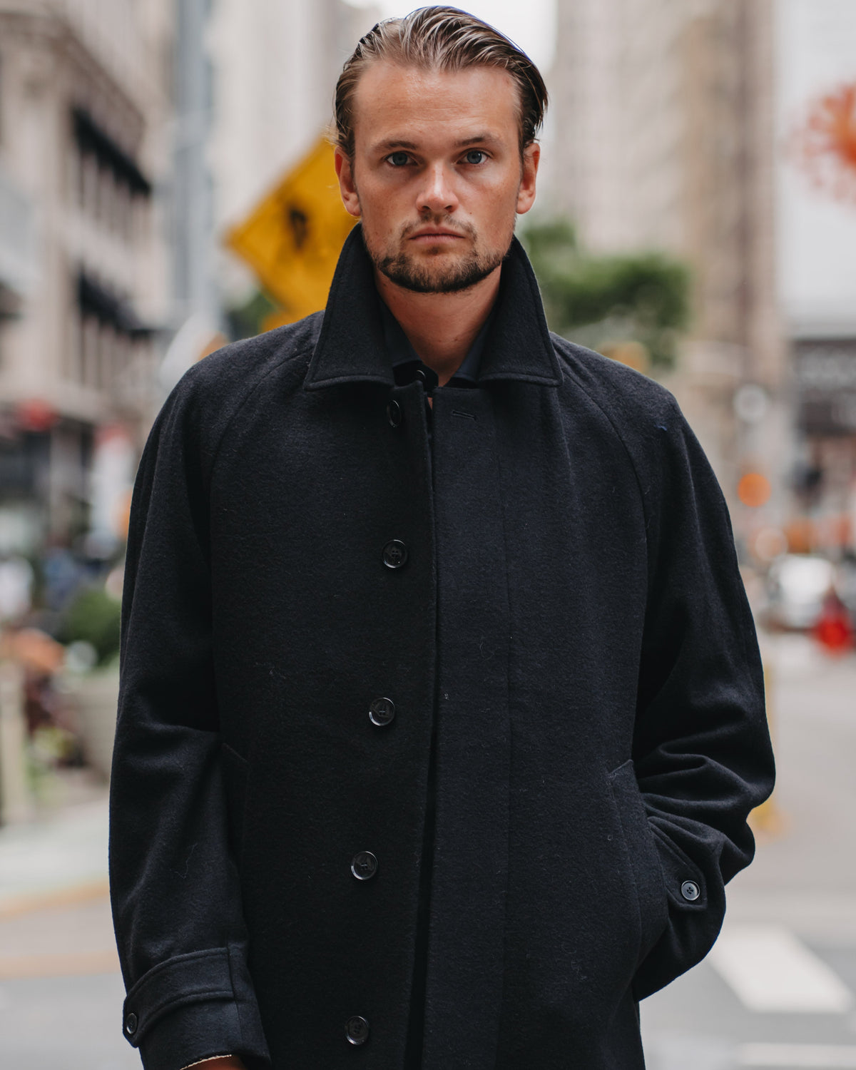 A man wearing a black wool-blend coat with a high collar and front buttons, standing on a city street with blurred buildings in the background.