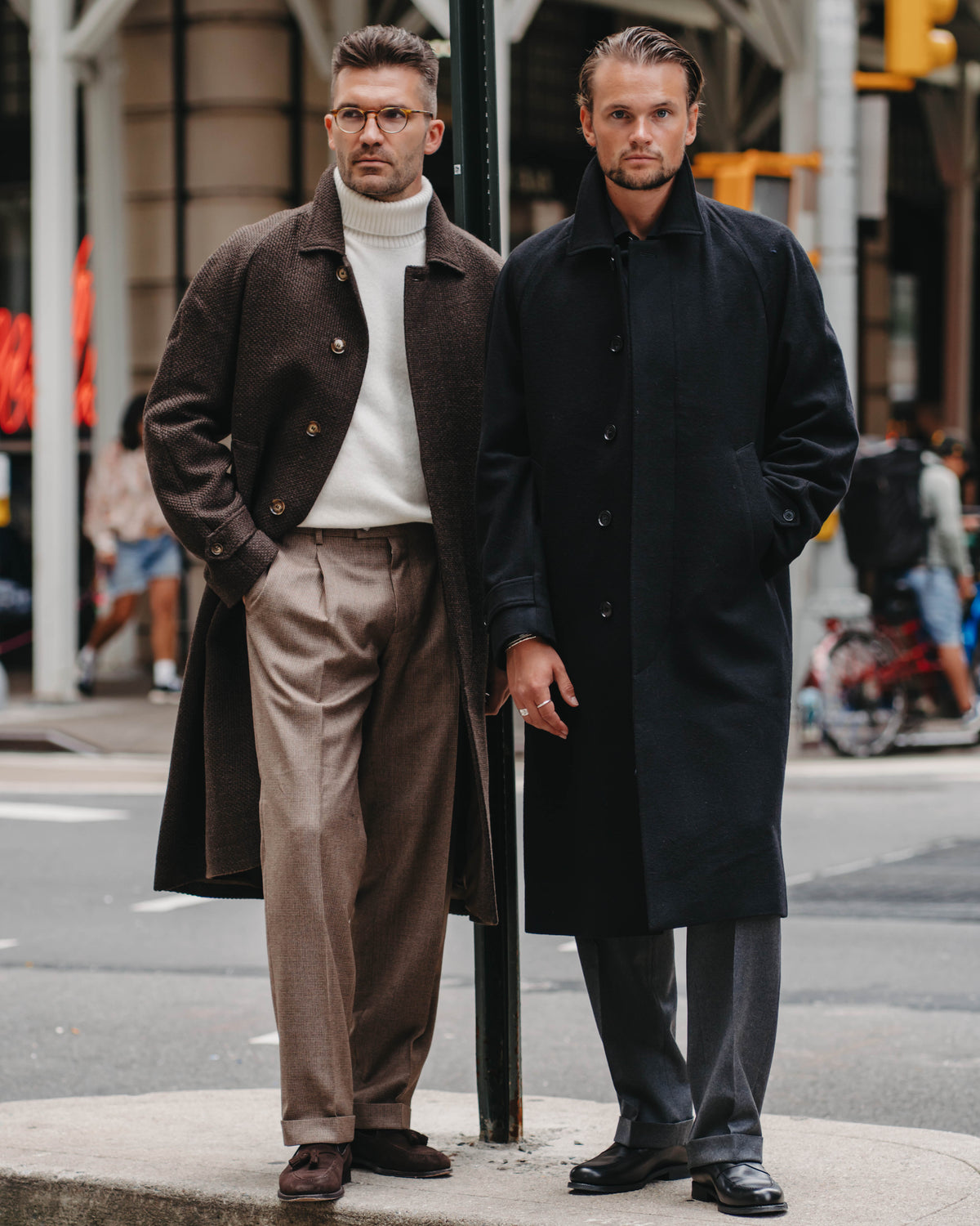 Two men standing on a city sidewalk next to a street sign, wearing long overcoats. The man on the left wears a brown coat over a white turtleneck and beige trousers, while the man on the right wears a black coat with gray trousers.