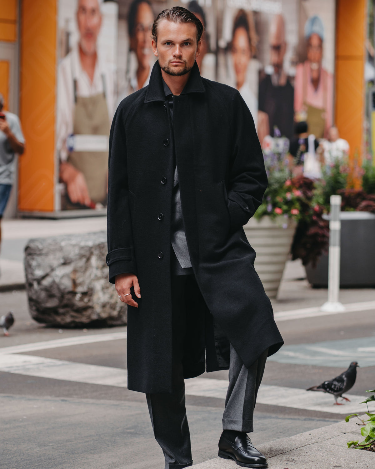 A man wearing a long black overcoat, dark shirt, gray trousers, and black shoes stands on a city sidewalk in front of a storefront with orange columns and a large outdoor advertisement.