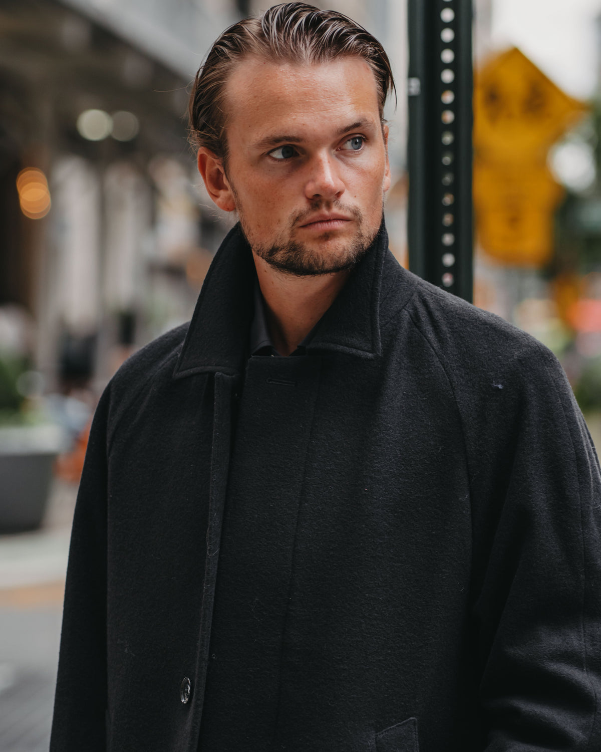 A man wearing a black wool coat stands outdoors on a city street, looking slightly off-camera. The background is softly blurred with urban elements like a pole and streetlights.