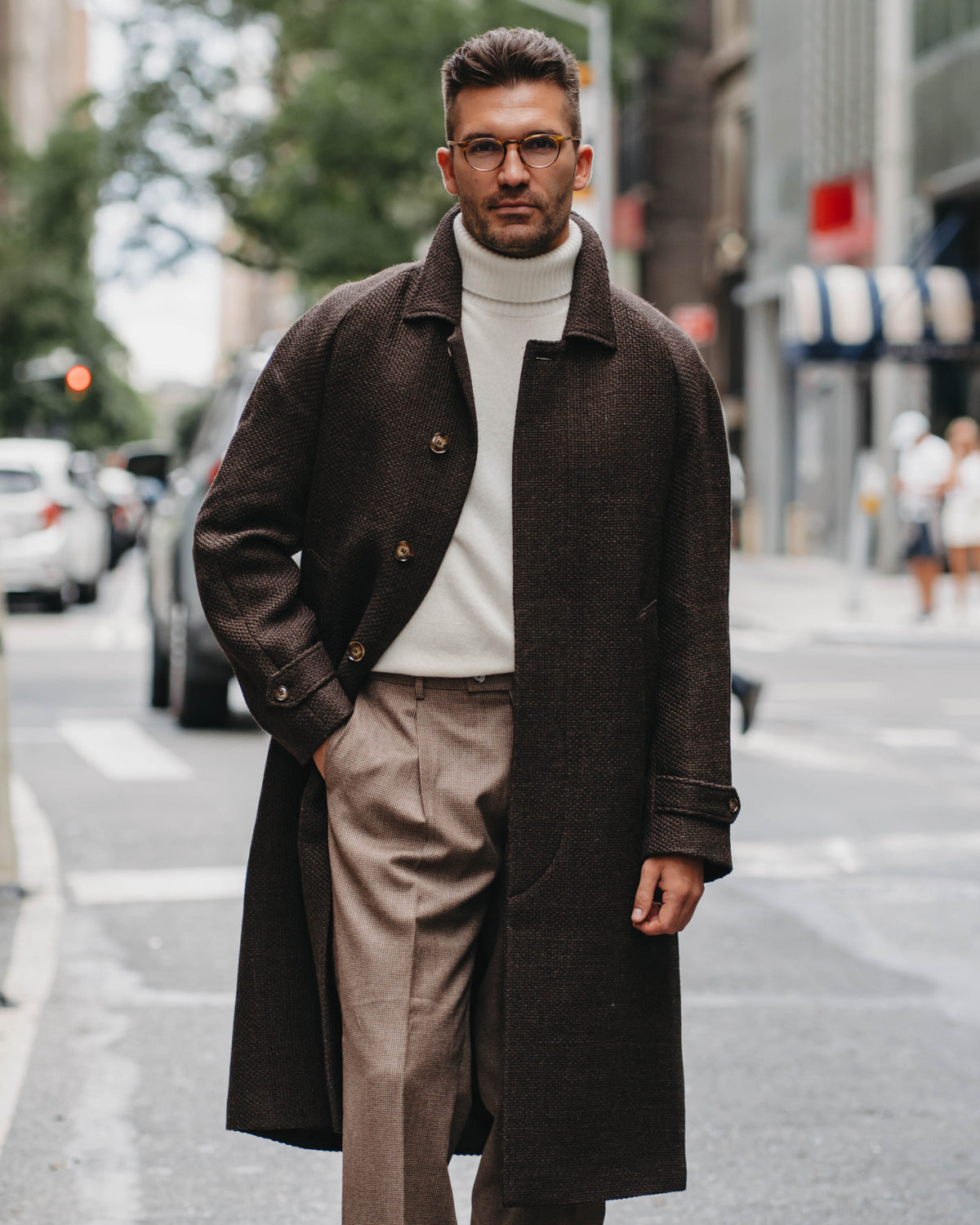 A man wearing a dark brown double-breasted overcoat, cream turtleneck sweater, beige trousers, and glasses walking on a city street.