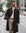 A man wearing a dark brown double-breasted overcoat, cream turtleneck sweater, beige trousers, and glasses walking on a city street.