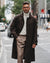 A man wearing a dark brown double-breasted overcoat, cream turtleneck sweater, beige trousers, and glasses walking on a city street.