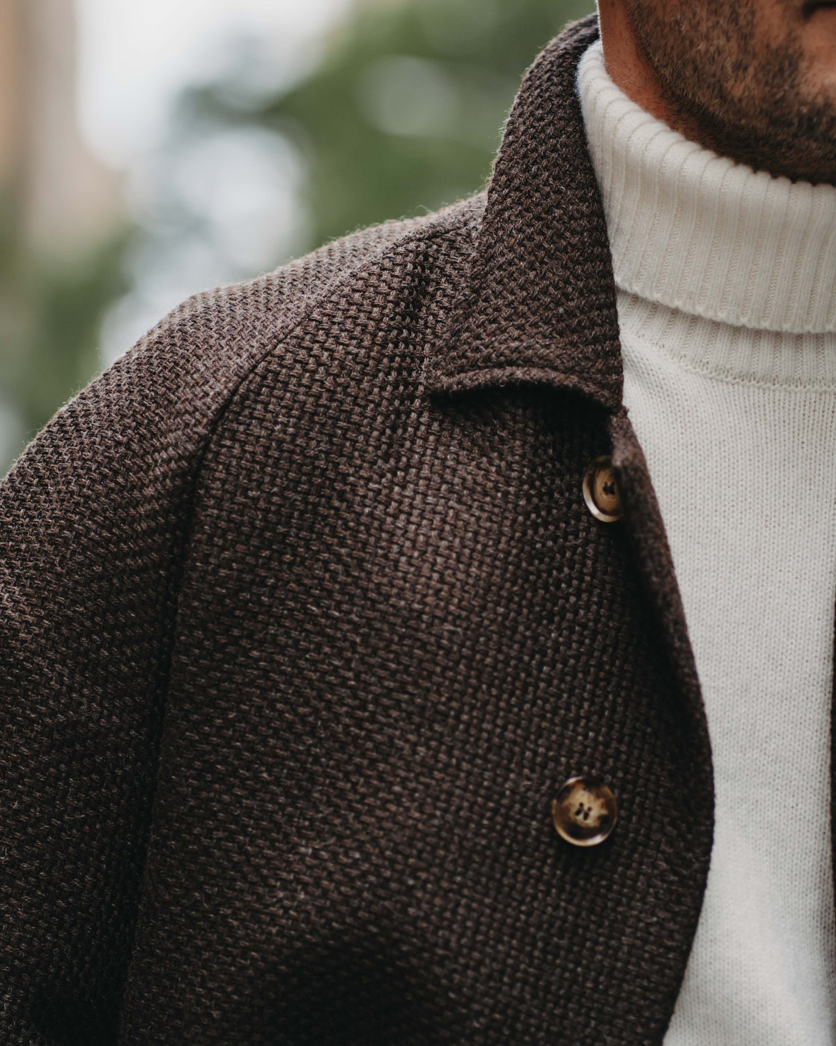 Close-up of a man wearing a dark brown textured wool-blend jacket over a white ribbed turtleneck sweater, with a softly blurred outdoor background.