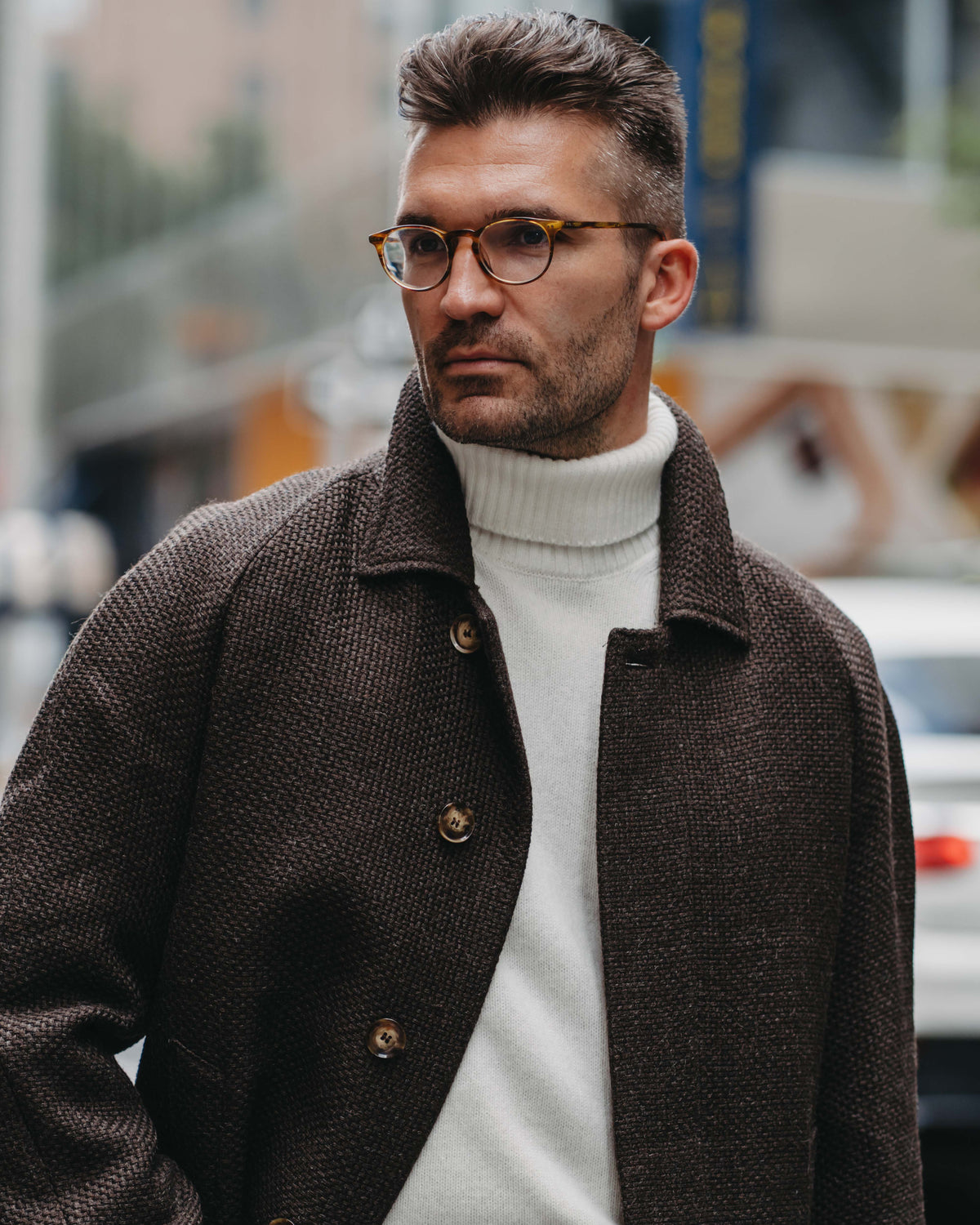 A man wearing a dark brown textured tweed jacket over a white turtleneck sweater, with tortoiseshell glasses, standing outdoors on a city street.