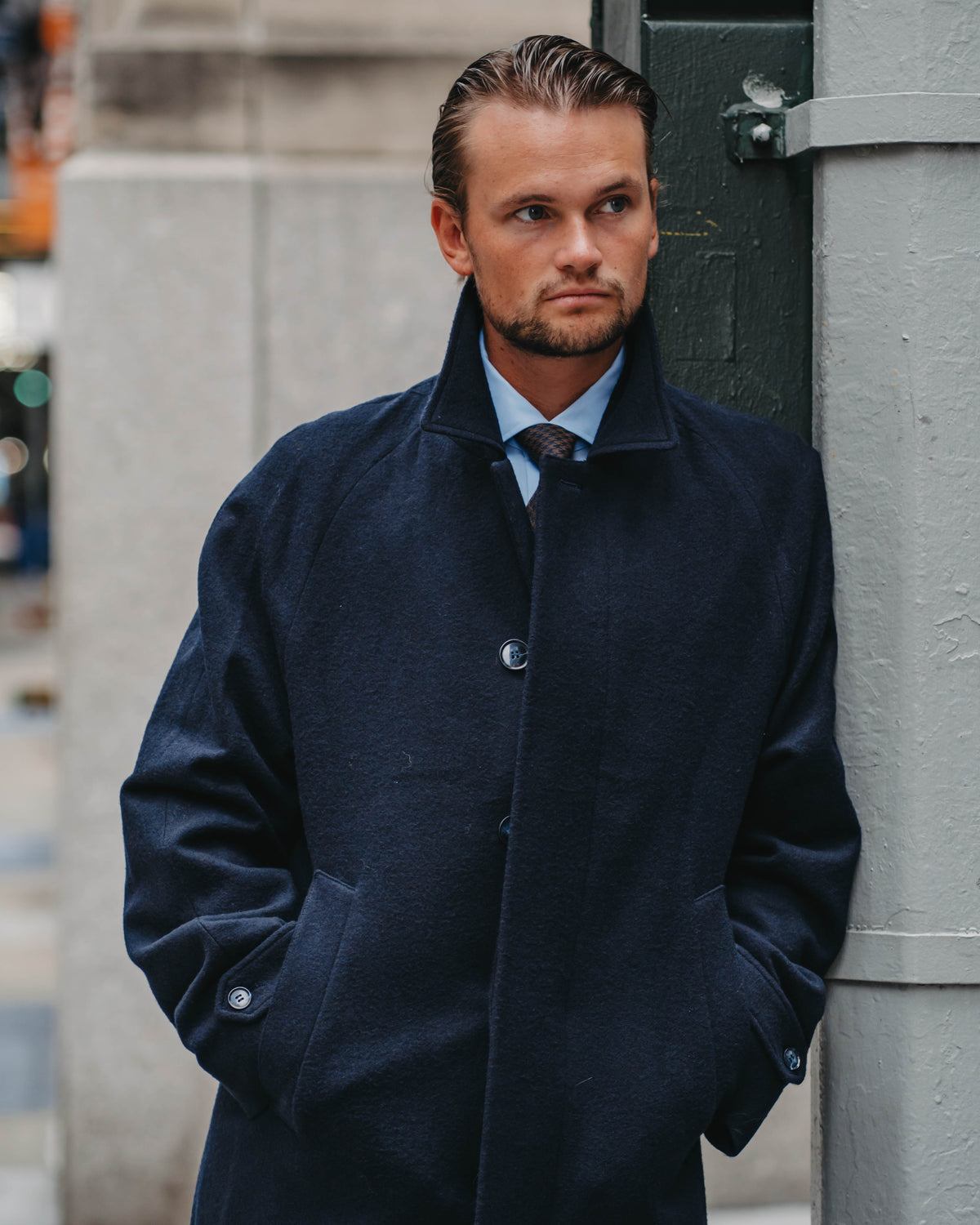A man wearing a dark navy blue overcoat over a light blue dress shirt and dark tie, standing outdoors next to a gray pole.