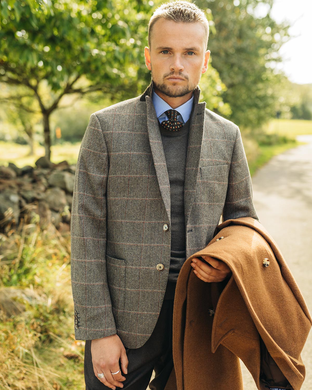 Man in a brown checkered blazer holding a brown coat outdoors