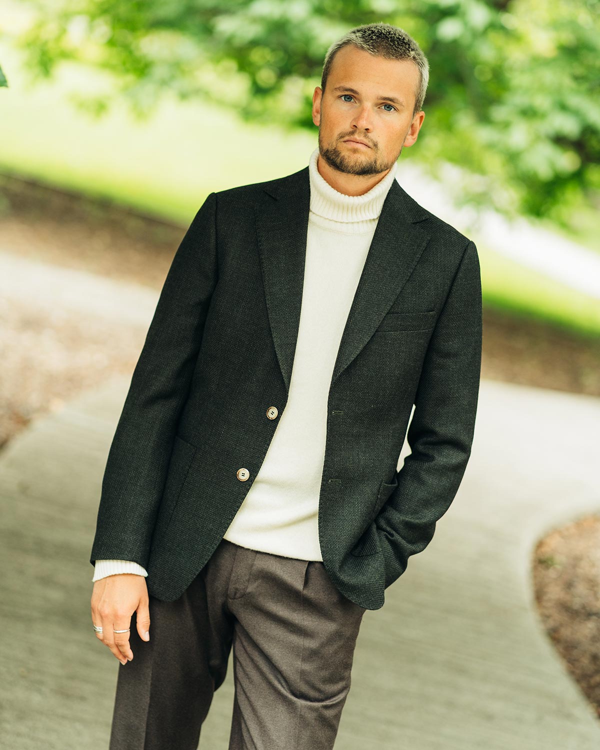 Man wearing a green blazer over a white rollneck sweater with a blurred green background