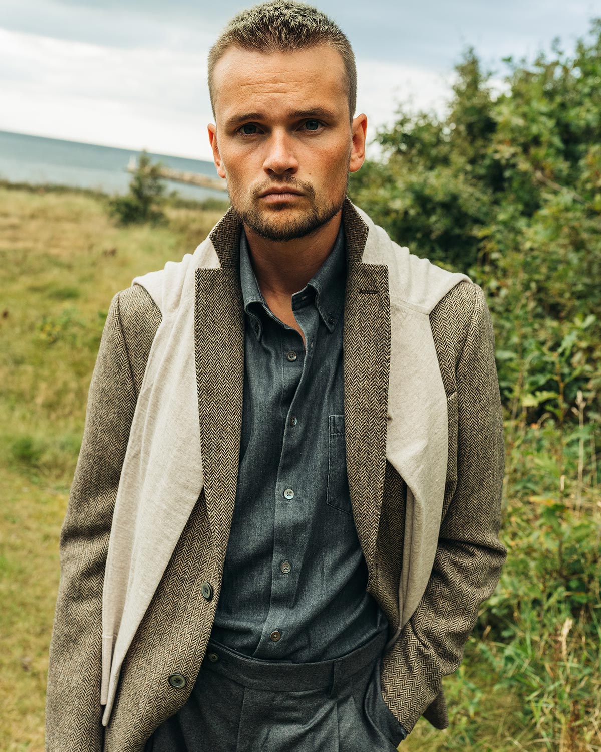 Man wearing a brown herringbone tweed jacket with a sweater on the shoulders in a natural setting