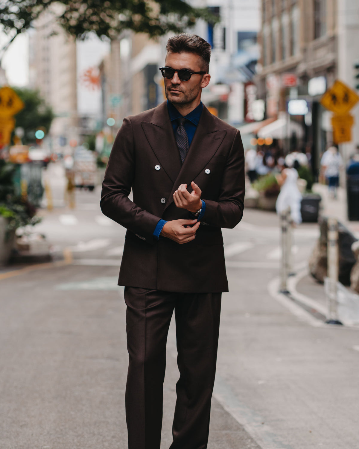 A man wearing a dark brown double-breasted suit, a blue shirt, and sunglasses stands on a city street.