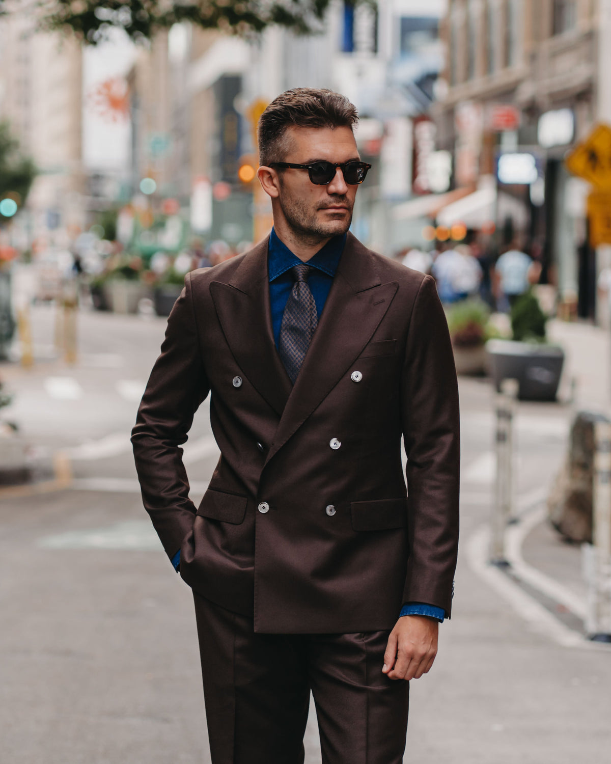 A man wearing a dark brown double-breasted suit, blue dress shirt, and dark tie walks down a city street wearing sunglasses.