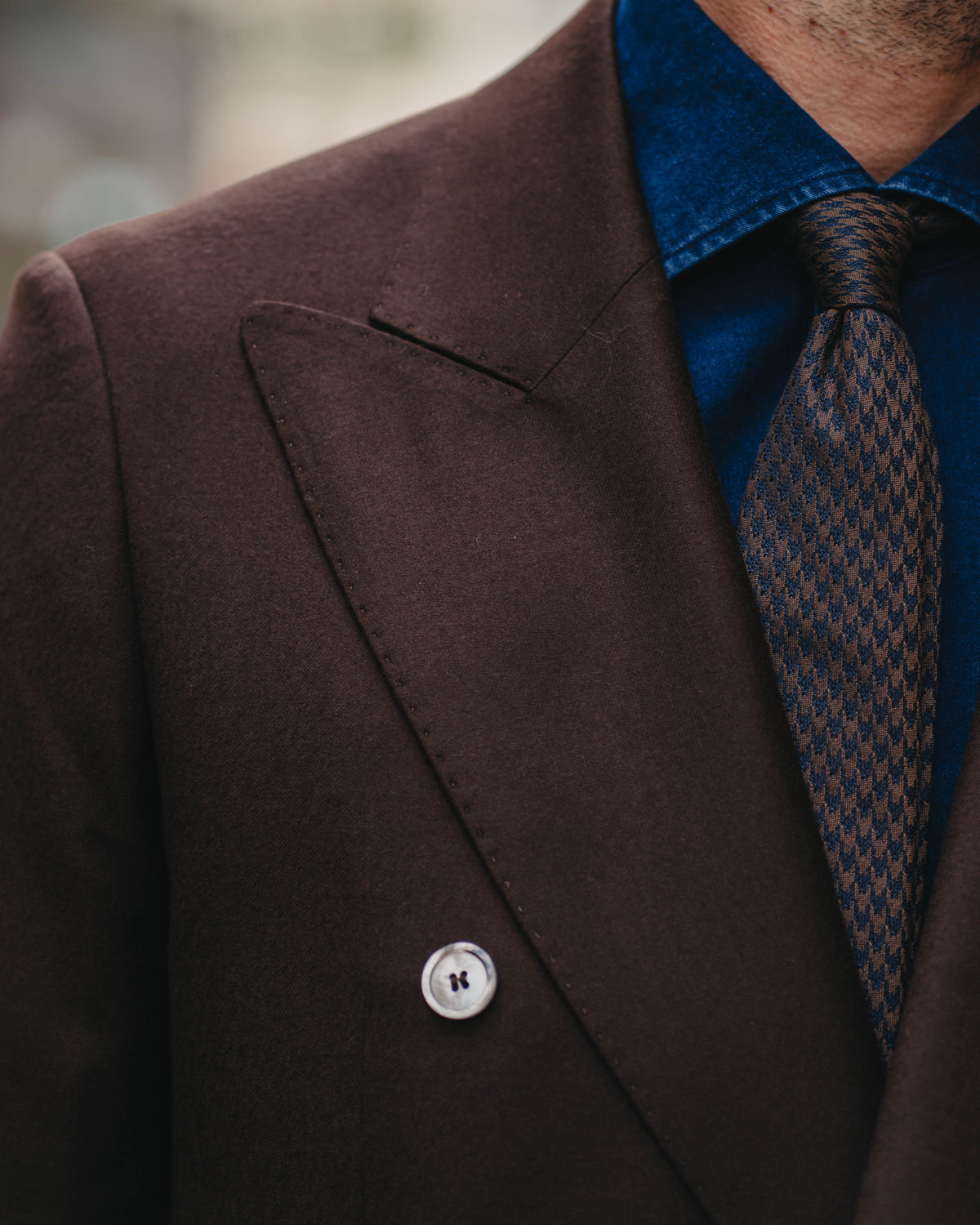 Close-up of a man wearing a dark brown double-breasted suit jacket, a textured blue shirt, and a patterned brown and blue tie.
