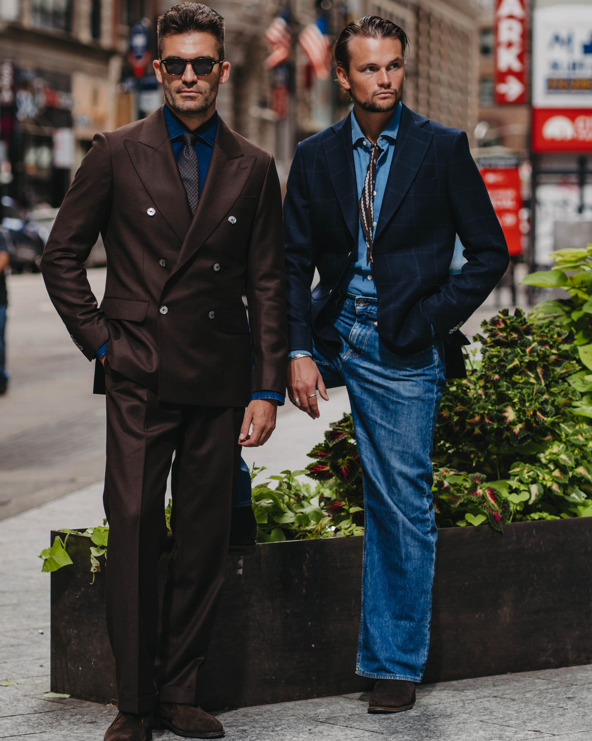 Two men standing on a city street wearing tailored suits. The man on the left wears a dark brown double-breasted suit with a dark blue shirt and tie.