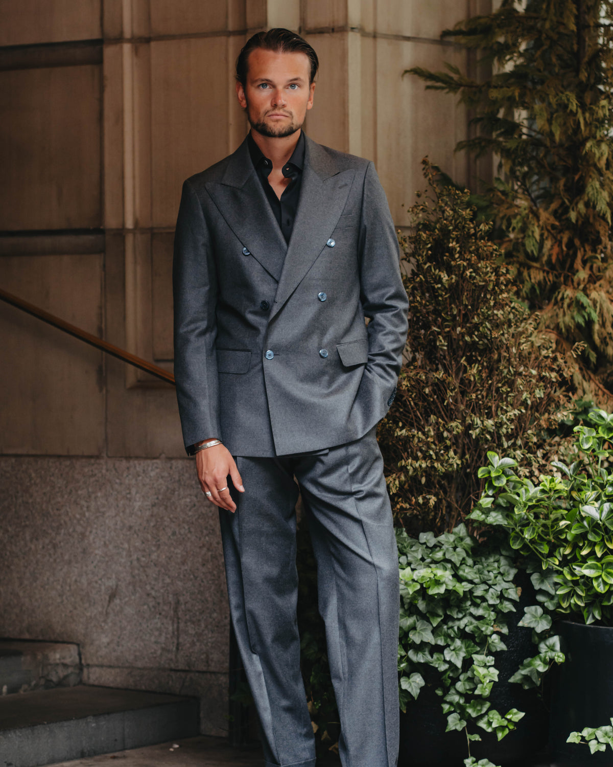 A man wearing a gray double-breasted suit with a black shirt and black dress shoes, standing outdoors against a stone building wall.