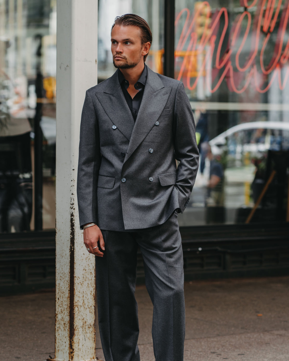 A man wearing a dark gray double-breasted suit with a black shirt and black dress shoes, standing on a city sidewalk in front of a storefront window.