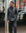 A man wearing a dark gray double-breasted suit with a black shirt and black dress shoes, standing on a city sidewalk in front of a storefront window.