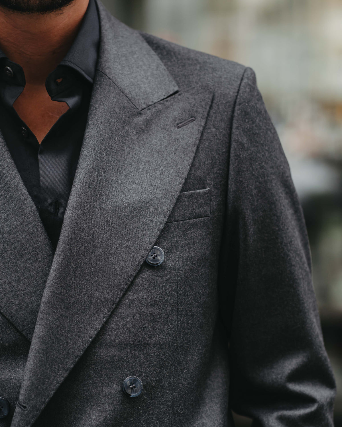 Close-up of a man wearing a dark gray double-breasted blazer over a black collared shirt, with a softly blurred outdoor background.