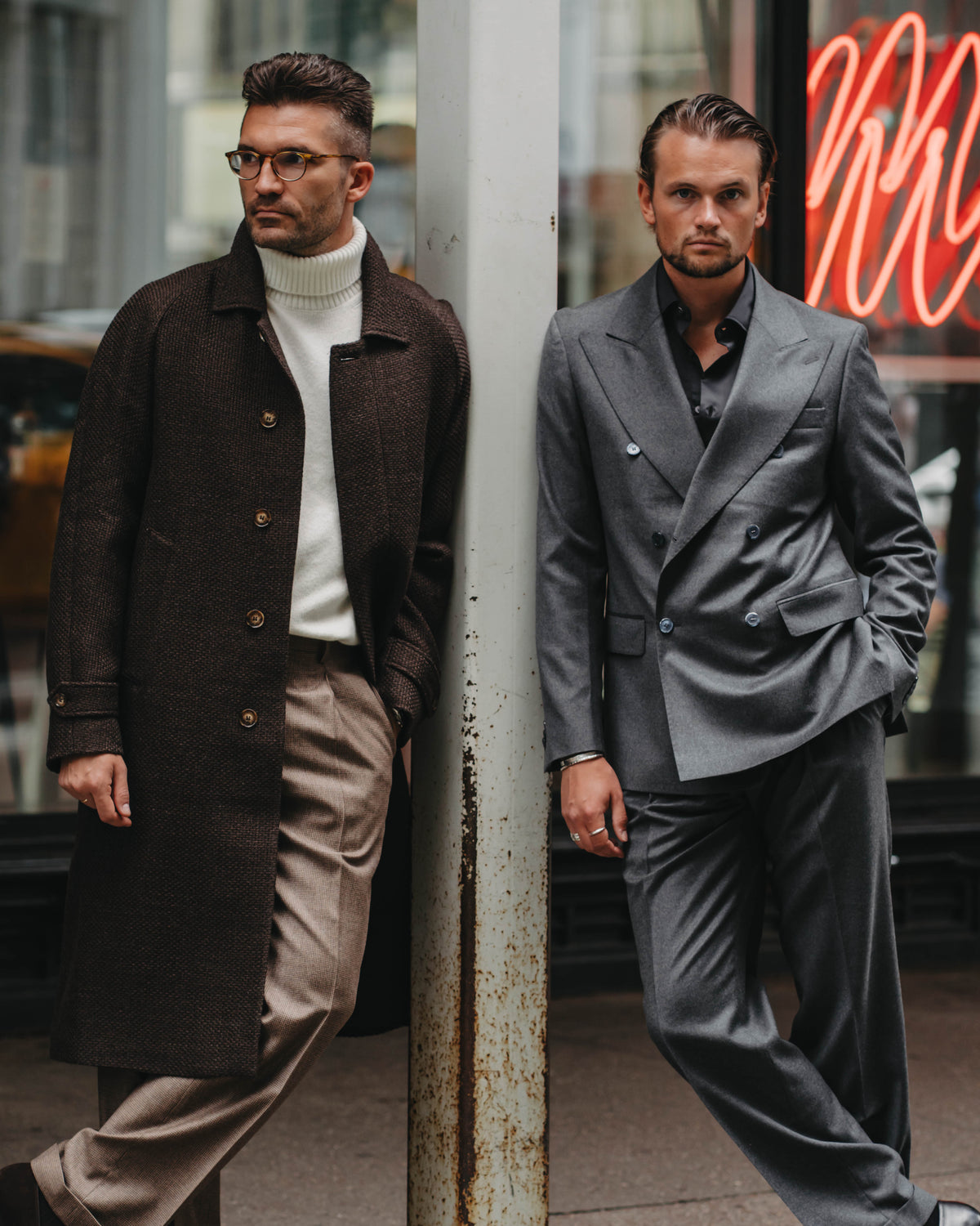 Two men standing on a city sidewalk beside a metal pole, dressed in formal menswear. The man on the left wears a dark brown overcoat over a white turtleneck and beige trousers.
