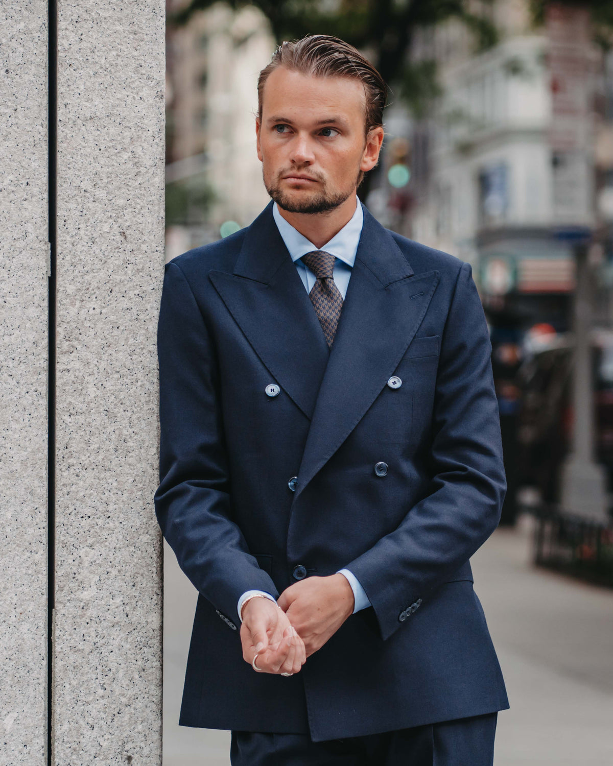 A man wearing a navy blue double-breasted suit, light blue dress shirt, and patterned tie stands against a gray stone pillar on a city street.