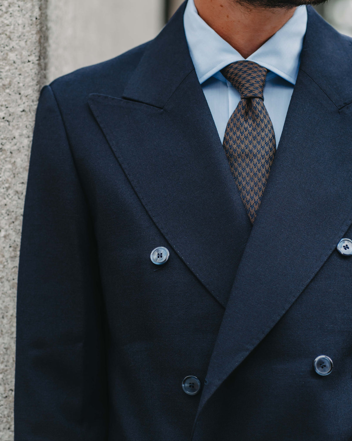 A close-up of a man wearing a dark navy double-breasted suit jacket, a light blue dress shirt, and a brown patterned tie, standing against a textured gray wall.