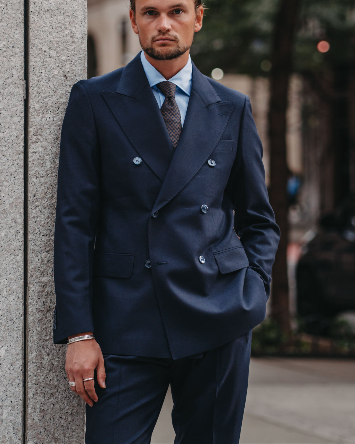 A man wearing a navy blue double-breasted suit, light blue dress shirt, and patterned tie, standing outdoors next to a stone pillar.