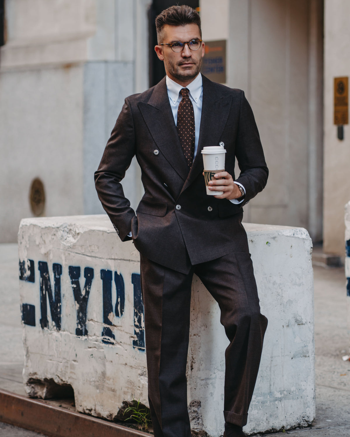 A man wearing a dark brown double-breasted suit, white dress shirt, and a brown patterned tie stands outdoors leaning against a concrete barrier, holding a takeaway coffee cup.