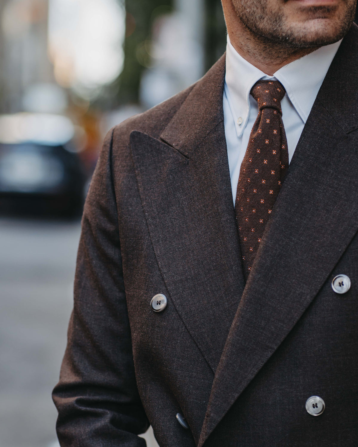 A close-up of a man wearing a dark brown double-breasted suit jacket, a white dress shirt, and a brown patterned tie, set against a blurred city street background.