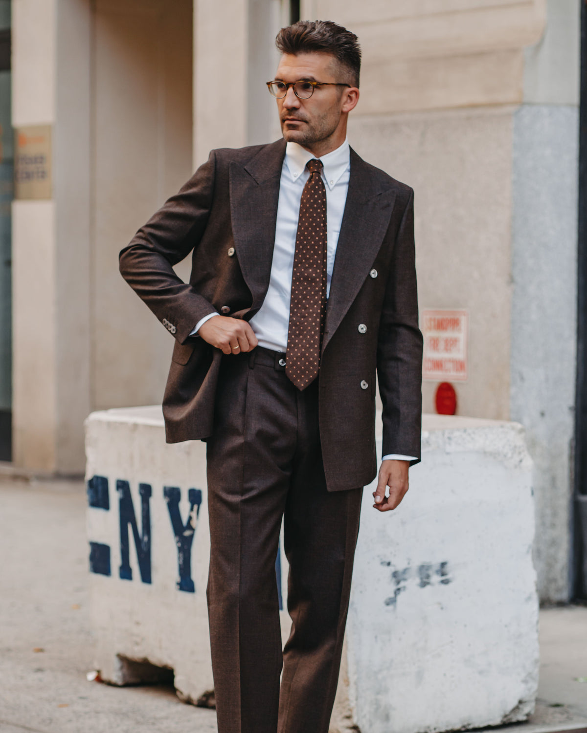 A man wearing a dark brown double-breasted suit, white dress shirt, and brown patterned tie stands outdoors on a city sidewalk.
