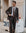 A man wearing a dark brown double-breasted suit, white dress shirt, and brown patterned tie stands outdoors on a city sidewalk.