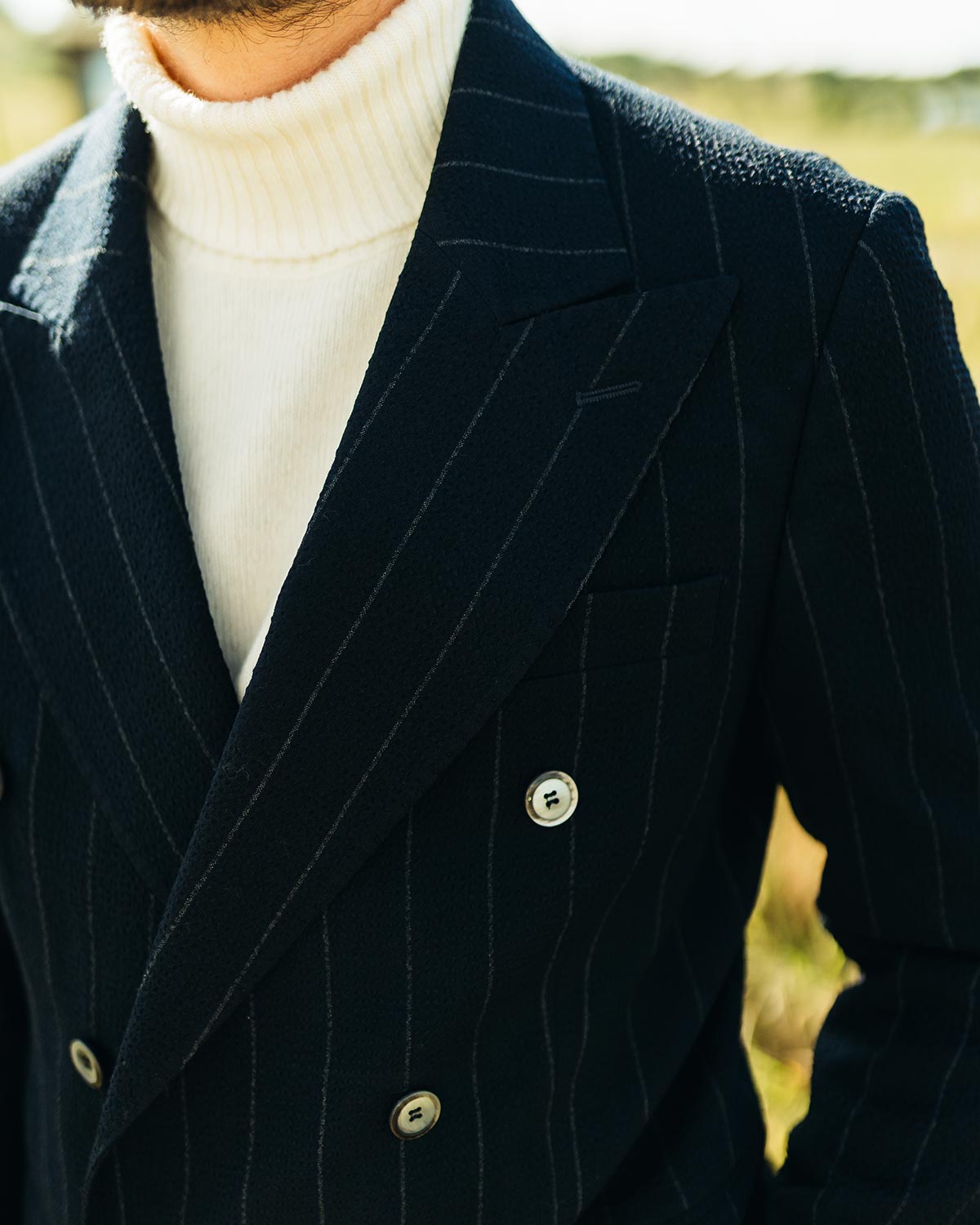 Navy pinstripe seersucker suit jacket with white turtleneck sweater against a blurred natural background