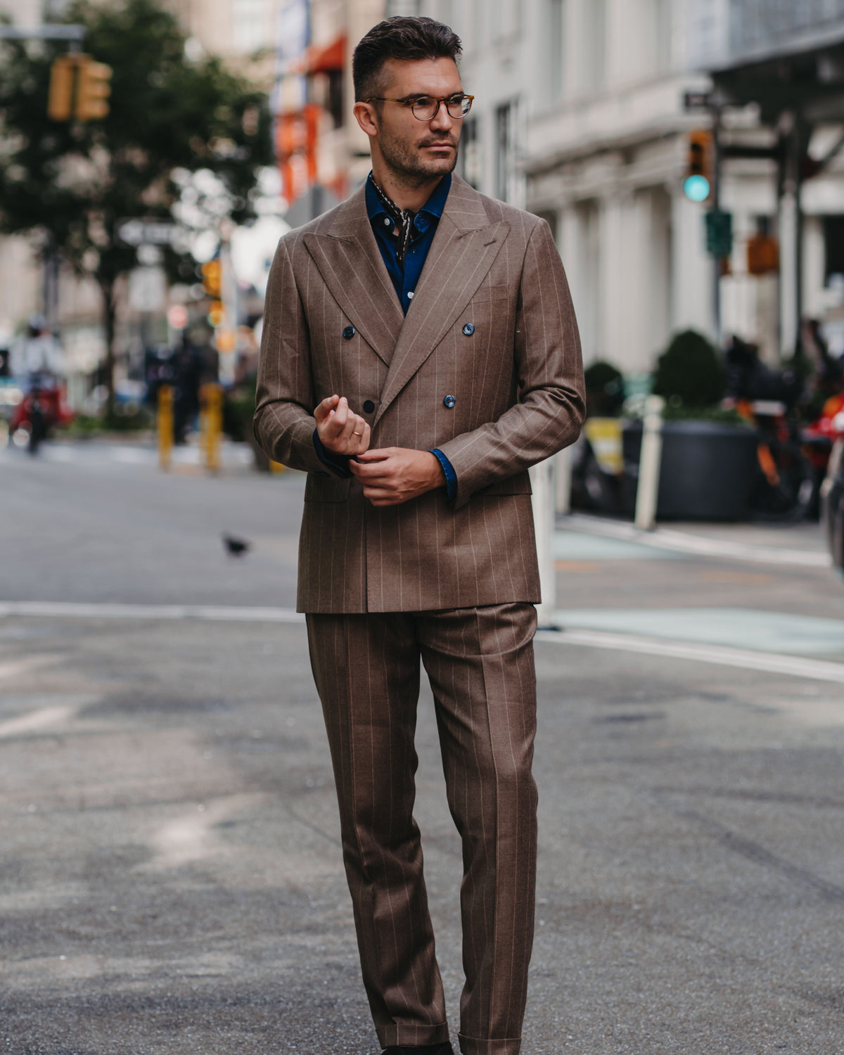 A man wearing a brown double-breasted pinstripe suit with a blue shirt and glasses, standing on a city street.