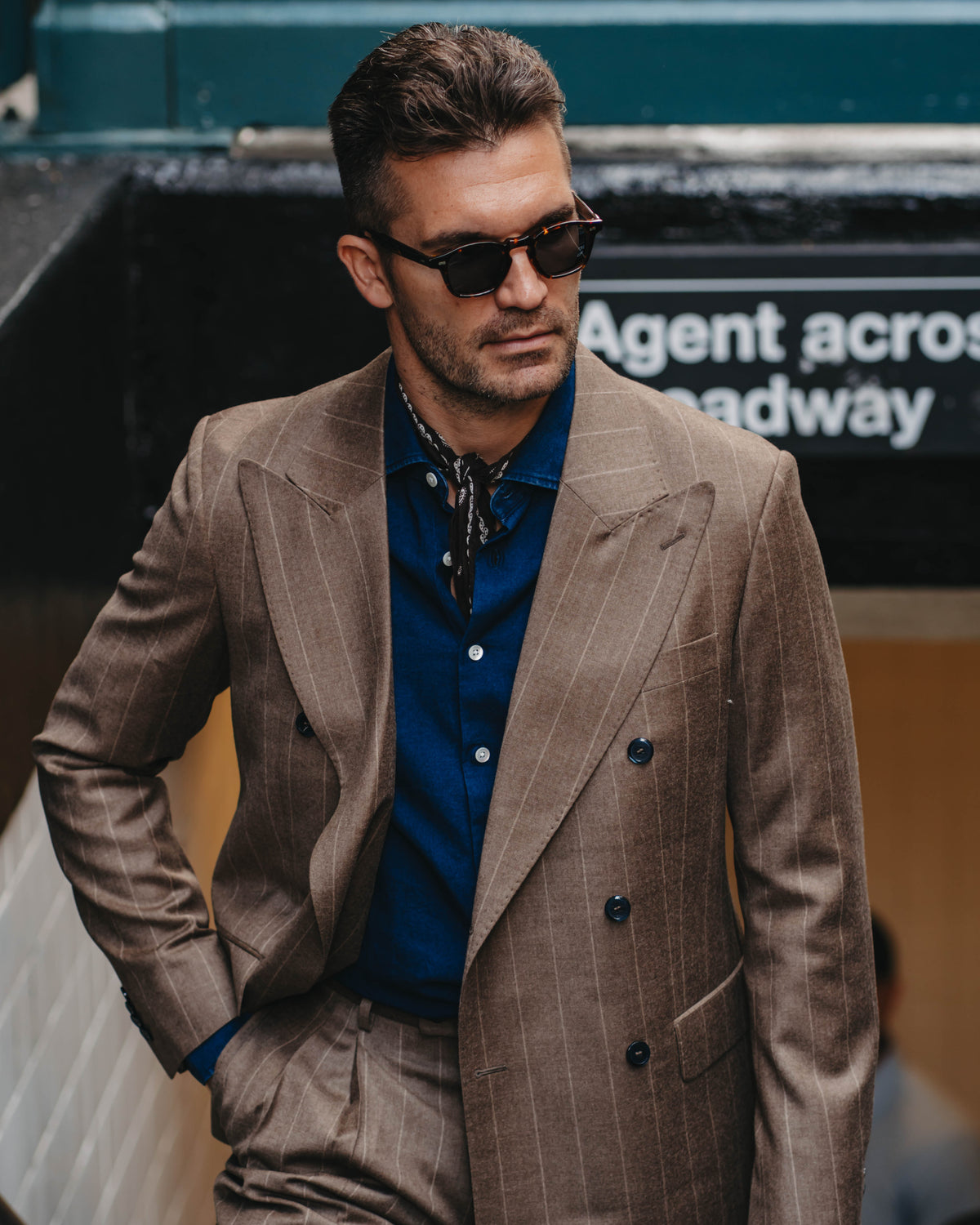 A man wearing a brown pinstripe double-breasted suit jacket and matching trousers, styled with a blue button-down shirt and dark sunglasses, standing outdoors.