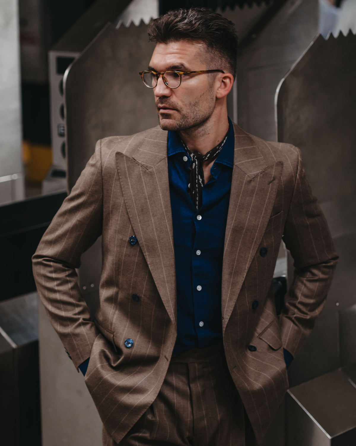 A man wearing a brown double-breasted pinstripe suit with a blue shirt and glasses, standing in front of metallic industrial machinery.