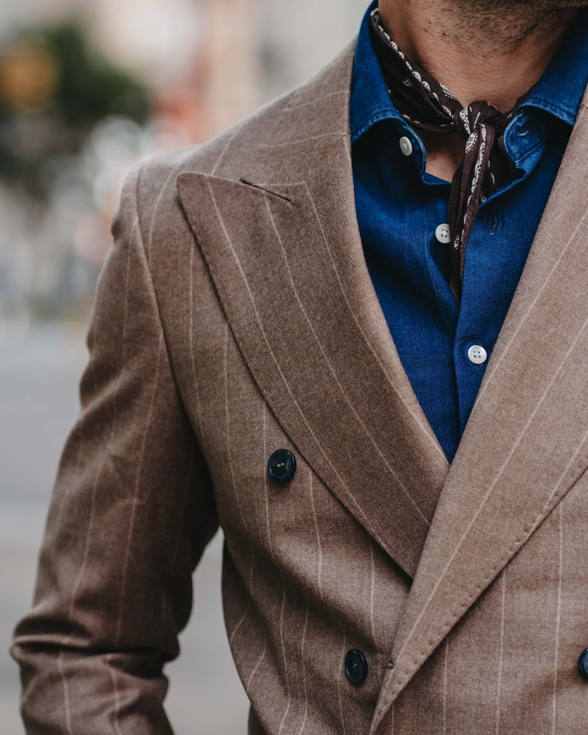 A close-up of a man wearing a brown double-breasted pinstripe blazer over a blue button-down shirt with a dark brown necktie.