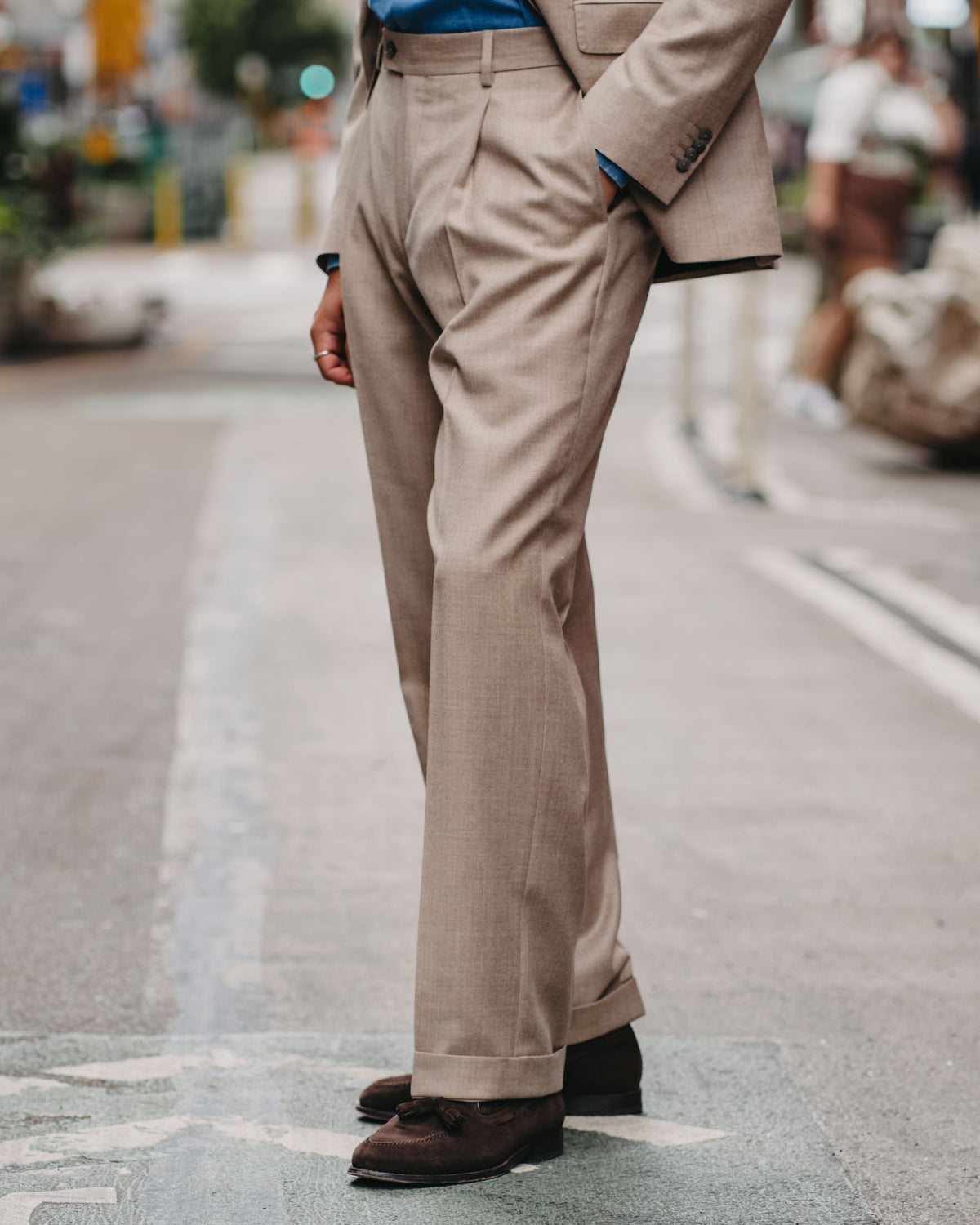 A man wearing a beige suit with a blue button-down shirt and dark brown leather shoes, standing on a city street.