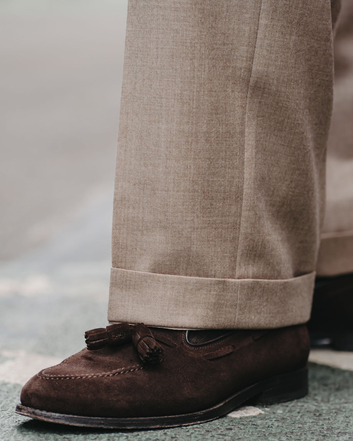 A close-up shot of a person's lower legs wearing beige trousers and a dark brown suede tassel loafer on a textured gray pavement.