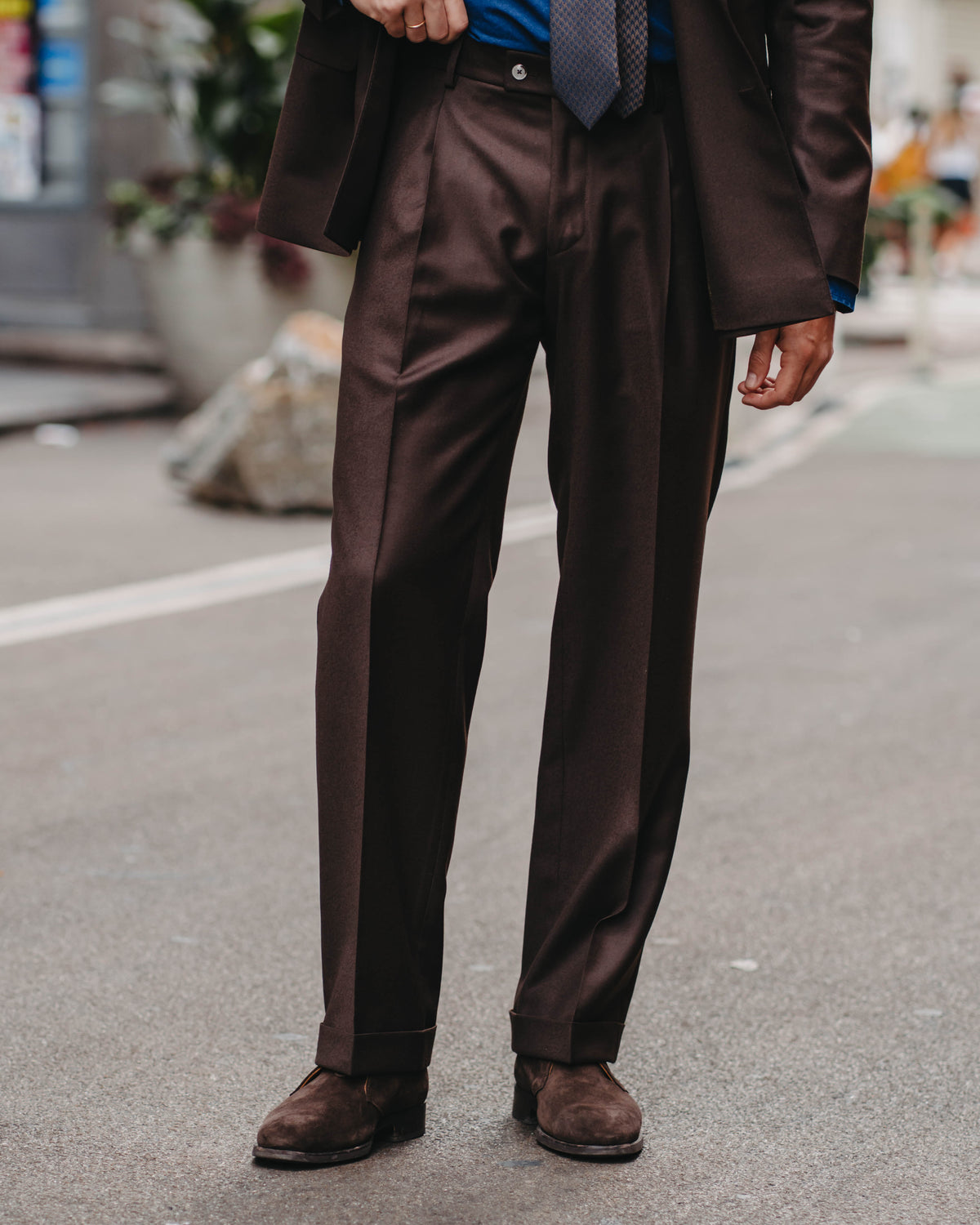 A man wearing a dark brown suit with a blue shirt, a gray patterned tie, and brown suede shoes, standing on a city street.