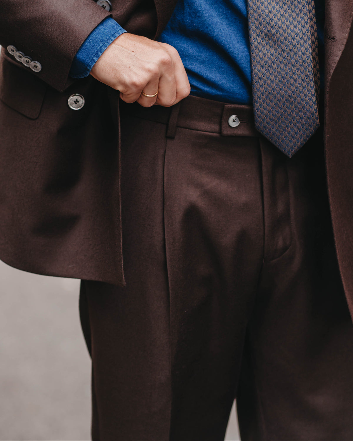 A close-up of a person wearing a dark brown suit, featuring a matching brown blazer and trousers, a blue shirt, and a patterned blue and gray tie.