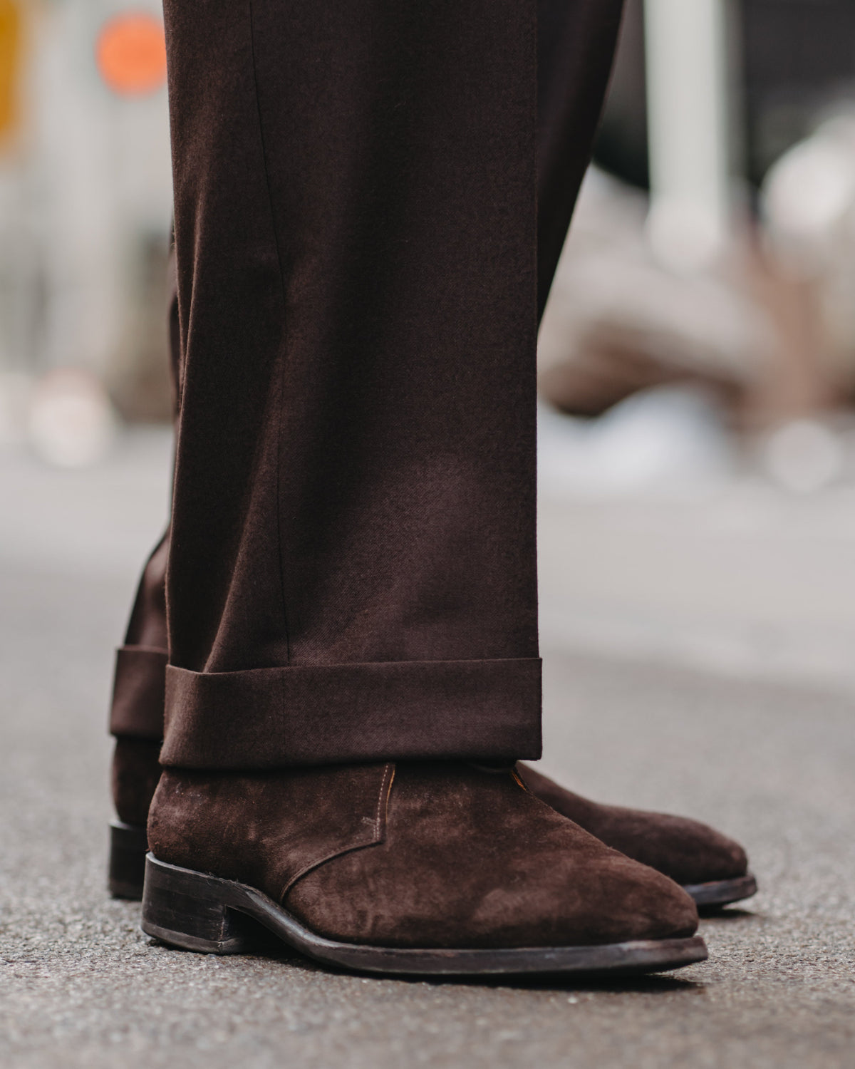 A close-up shot of a person's legs wearing dark brown suede boots and matching dark brown trousers, standing on a gray asphalt street.