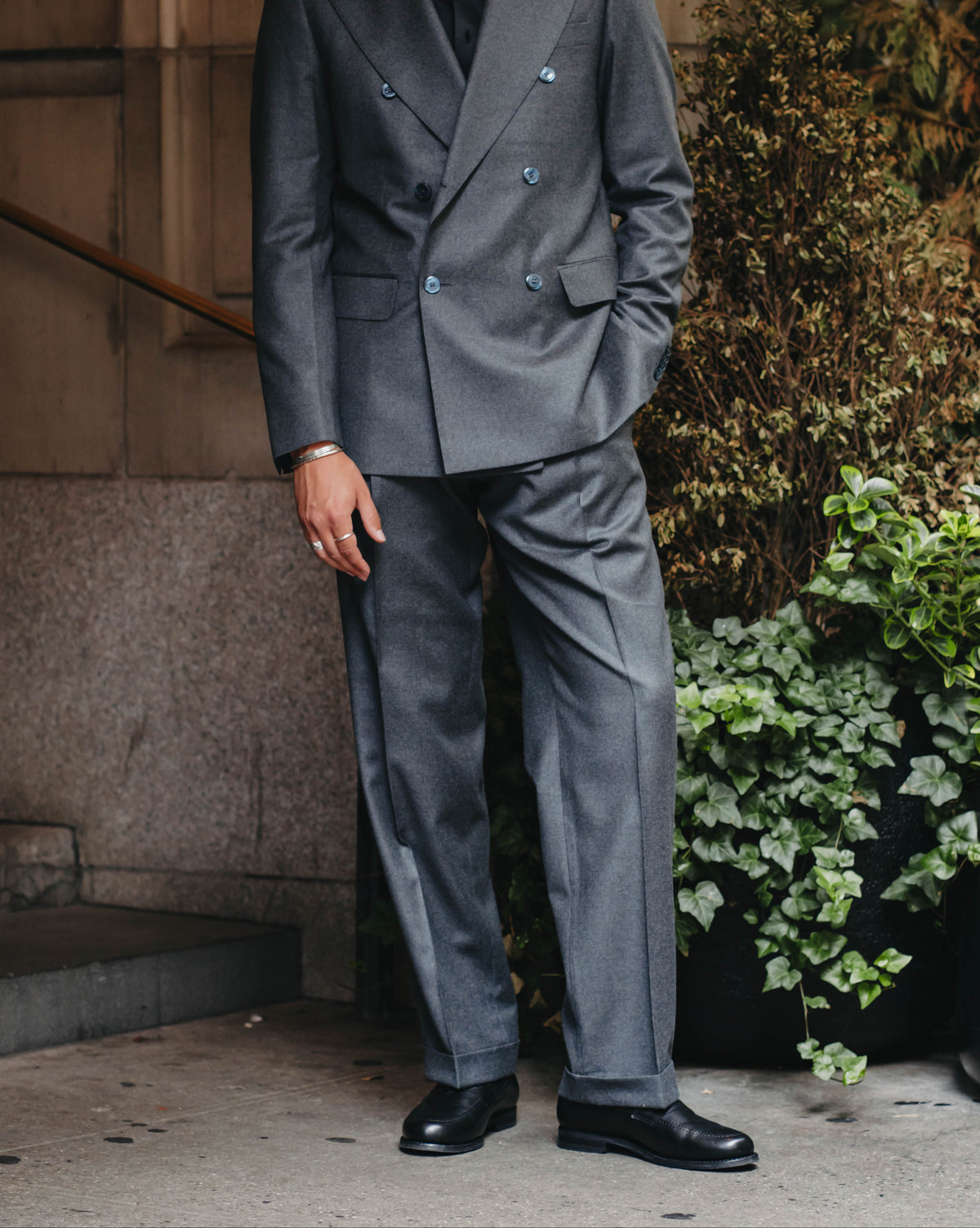 A man wearing a gray double-breasted suit with a black shirt and black dress shoes, standing outdoors against a stone building wall with green plants.