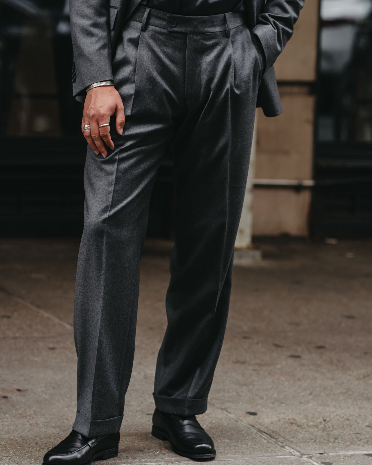 A man wearing a dark charcoal gray two-piece suit with a matching shirt and black dress shoes, standing on a city sidewalk.
