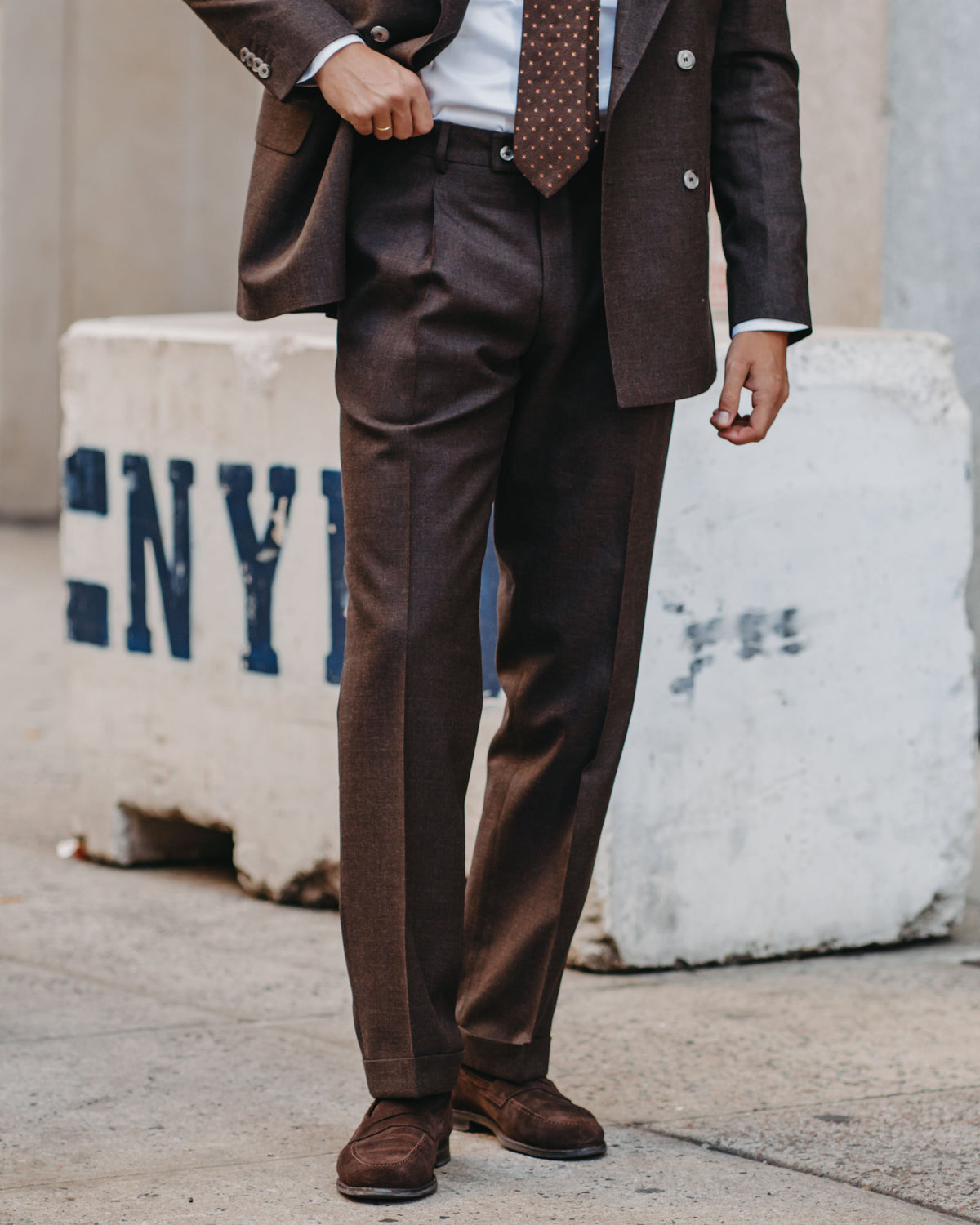 A man wearing a dark brown double-breasted suit with a white shirt and a brown patterned tie, standing outdoors in front of a concrete barrier.