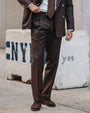 A man wearing a dark brown double-breasted suit with a white shirt and a brown patterned tie, standing outdoors in front of a concrete barrier.