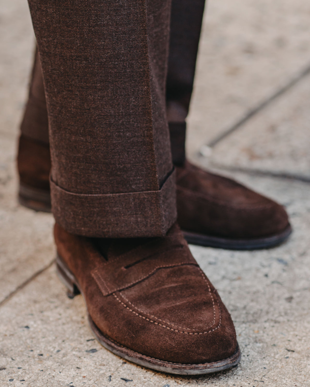 A close-up shot of a person's legs wearing dark brown suede penny loafers and matching brown trousers, standing on a light gray concrete pavement.