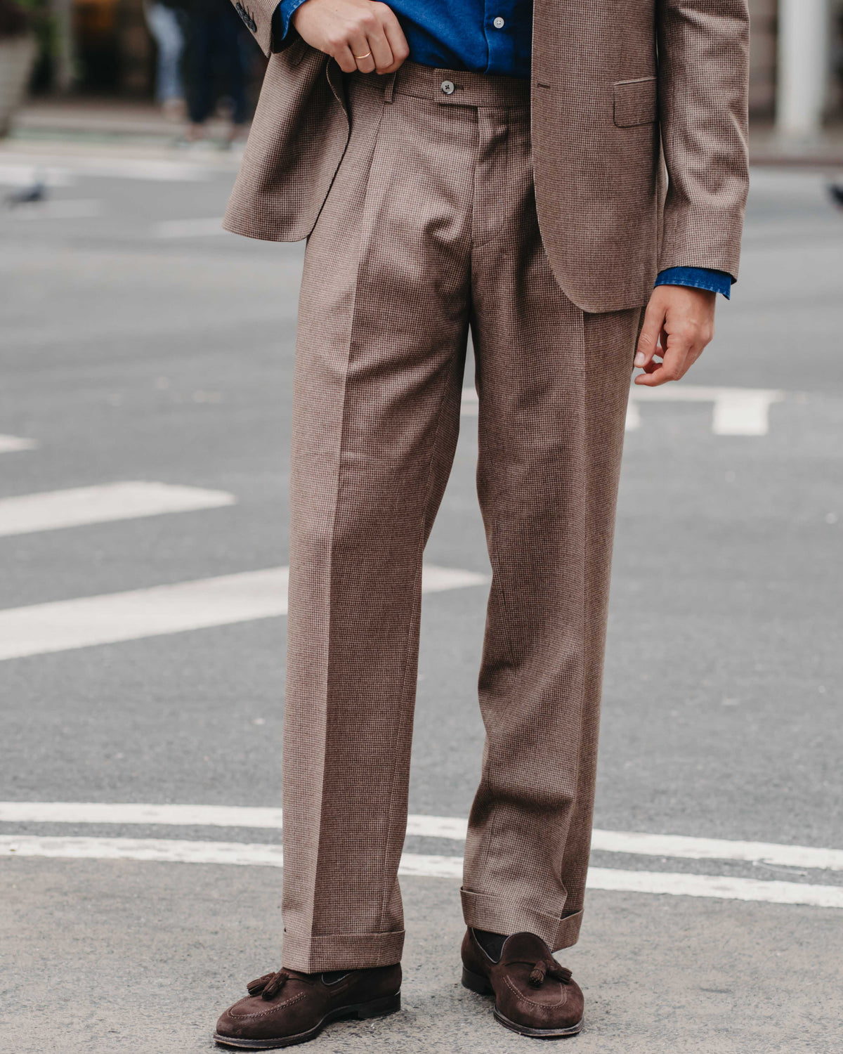 A man wearing a brown two-piece suit with a blue button-down shirt and dark brown suede loafers, standing on an urban street.