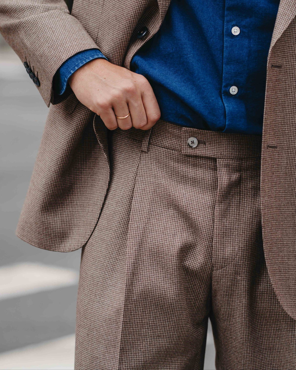 A close-up of a person wearing a brown and beige micro-check suit with a blue button-down shirt tucked inside, showing the hand adjusting the jacket.
