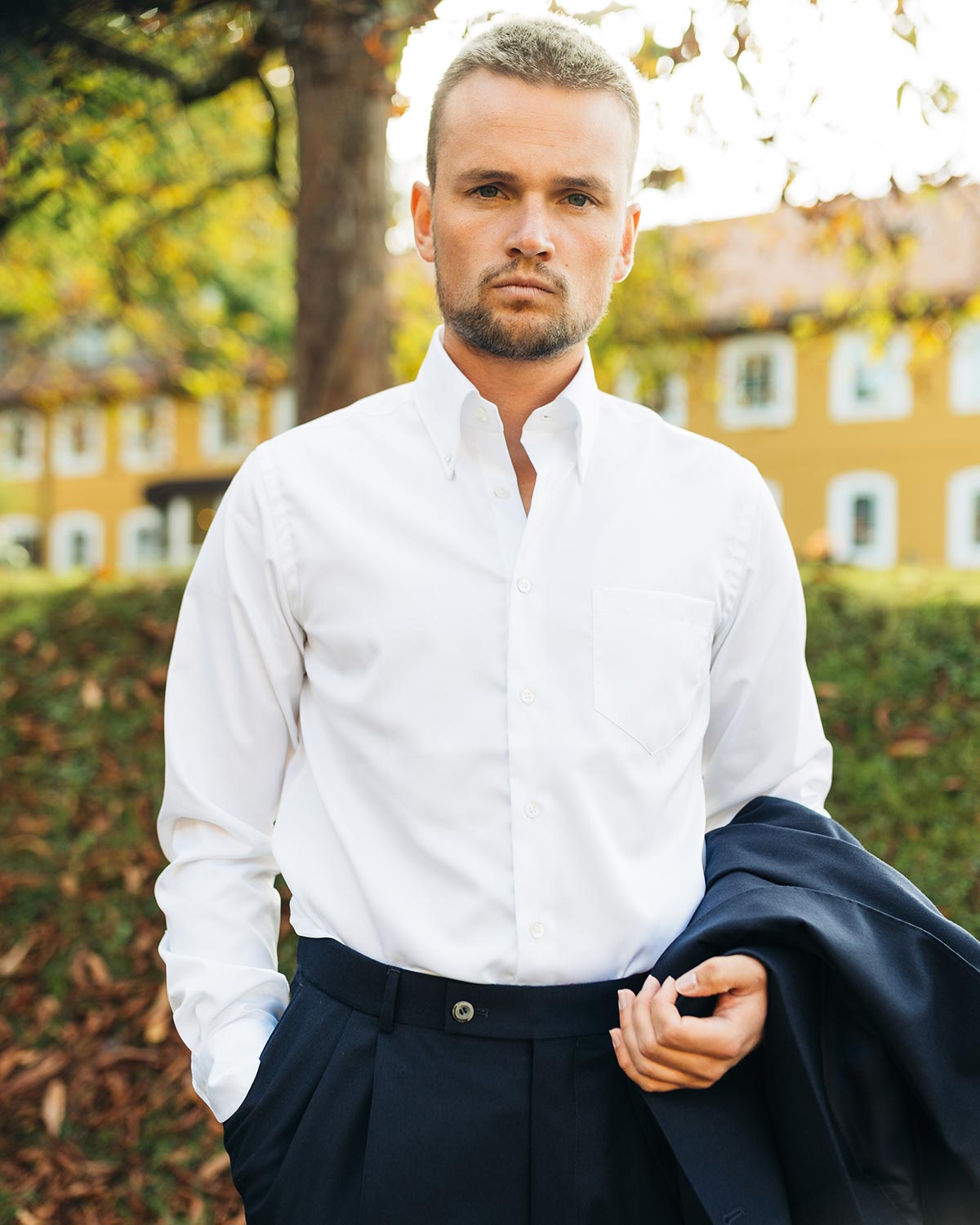 Man wearing a white dress shirt and dark pants outdoors with trees and buildings in the background
