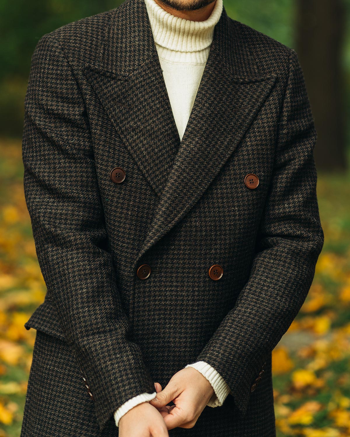 Person wearing a brown double breasted houndstooth patterned coat with a blurred natural background