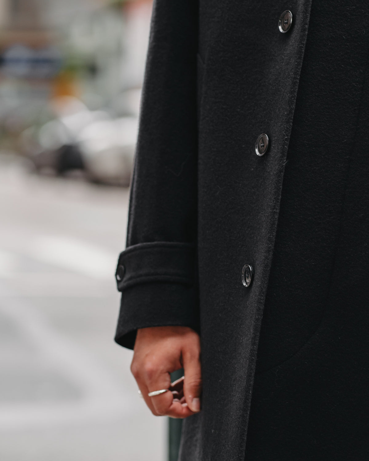 A close-up of a person wearing a long black coat outdoors. The coat has dark buttons and a dark cuff.